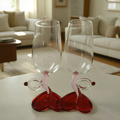 Two clear wine glasses with pink stems and red heart-shaped bases, displayed on a white table in a softly lit living room.