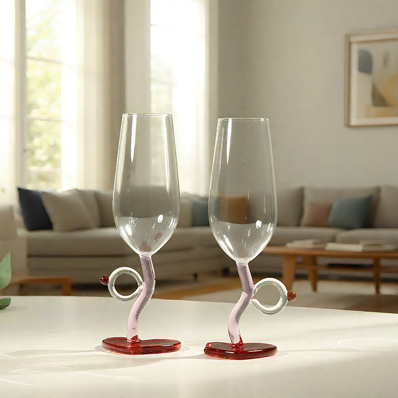 Two clear stemmed wine glasses with twisted pink bases and red circular feet, placed on a white table in a softly lit living room.