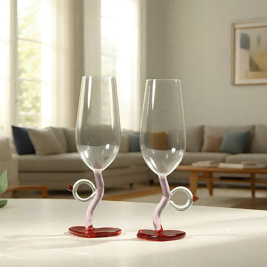 Two clear stemmed wine glasses with twisted pink bases and red circular feet, placed on a white table in a softly lit living room.