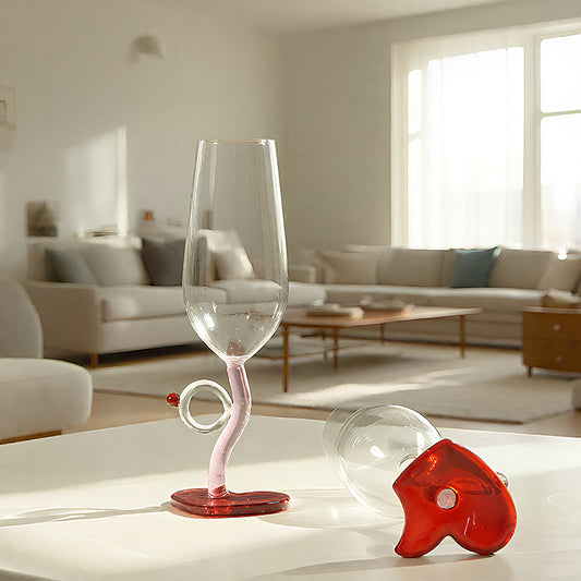 A clear stemmed wine glass with a pink curved stem and a small loop detail, shown beside a matching clear and red decorative base on a white table in a bright modern living room.