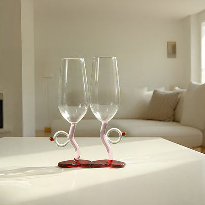 Two clear champagne flutes with pink and red twisted stem bases, displayed on a white table in a bright, modern living room.